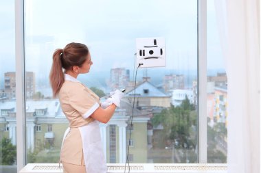 Üniformalı bir hizmetçi pencere temizleyici bir robotu çalıştırıyor. İç kısımda fotoğraflar var. Temizlik ve temizlik kavramı. Uzayı kopyala. Arka görünüm. Tanımlanamayan fotoğraf.
