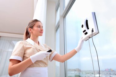 Üniformalı bir hizmetçi cama cam temizleyici bir robot yerleştiriyor. Pencereyi açın. İç kısımda fotoğraflar var. Temizlik ve temizlik kavramı. Alt manzara. Uzaktan kumanda hizmetçinin elinde..
