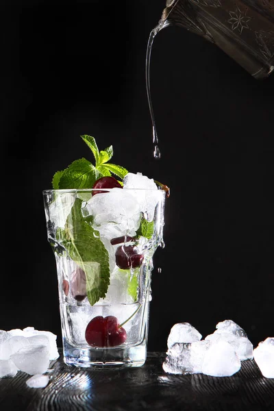 A limonada fresca é vertida de um vaso vintage em um copo com cerejas e gelo. Folhas de hortelã em um copo. Bebida sazonal de verão. Foto sobre um fundo preto. Cópia do espaço. Foto vertical . — Fotografia de Stock