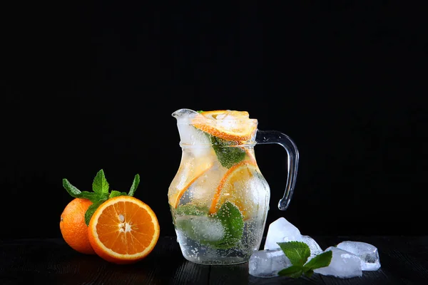 Limonada fresca feita de laranjas e hortelã em um jarro de vidro. Cubos de gelo na bebida. Foto sobre um fundo preto. Cópia do espaço . — Fotografia de Stock