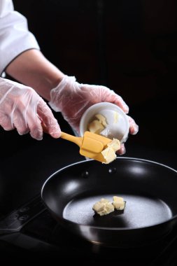 Aşçı tavaya bir spatulayla tereyağı ekler. Tereyağı dilimleri tavaya düşer. Tanımlanamayan dikey fotoğraf. Sadece eldivenli eller.