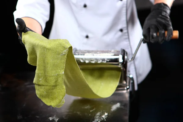 Homemade spinach pasta. A uniformed chef prepares pasta. Unrecognizable person. Photo on a black background.Machine for preparing pasta or a pasta machine. Copy space.