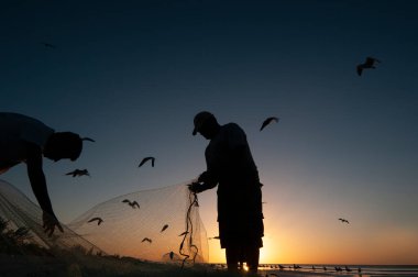 Sahilde gün batımında iki balıkçı arka plandaki balık ağlarını, renkli gökyüzünü ve Meksika 'daki Holbox Adası' ndaki martıları temizler.