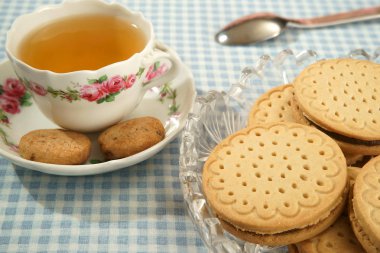Bitki çayıyla dolu porselen bardak. Çerezler çay tabağında ve kristal bir tabakta. Beyaz ve mavi Vichy masa örtüsü. Masada bir kaşık