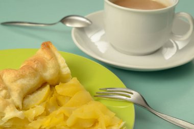 Bir parça tatlandırılmamış ev yapımı elma turtası yeşil bir tabakta tatlı çatalıyla. Sıcak çikolatayla dolu beyaz bir fincan. Kaşık ve çay tabağı. Eşyalar yeşil bir masaya yerleştirildi.