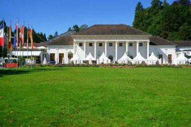 Kurhaus adında Baden-Baden Kumarhanesi. Restoran ve soldaki uluslararası bayraklar. Beyaz çadırlı bir etkinlik için devam eden hazırlıklar. Yeşil çimen ve mavi gökyüzü. Baden-Wuerttemberg, Almanya. Boşluğu kopyala.