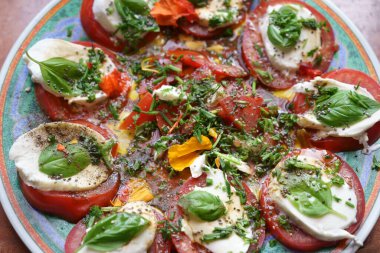 İçinde Mozzarella domates salatası olan bir tabağa yakından bak. Fesleğen yaprağı, soğan ve yenilebilir çiçek yapraklarıyla tatlandırılmış. Sos olarak zeytinyağı ve limon..