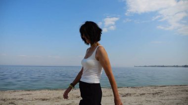 Güneşli bir günde sahilde kumsala giden mutlu yetişkin kadın. Arka planda sakin dalgalarla deniz kıyısında yürüyen güzel bir bayan. Yaz tatili ya da tatil kavramı. Yavaş çekim
