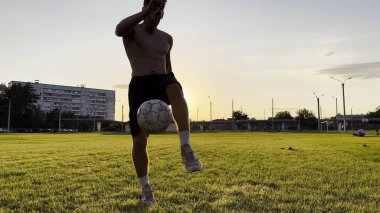 Genç adam gün batımında stadyumda futbol topuyla hokkabazlık yapıyor. Yeşil sahada profesyonel futbolcu topa vuruyor. Sporcu arka planda güneş ışığıyla çayırda numaralar yapıyor. Serbest stil futbol.