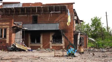 Ukrainian flag wave on flagpole while wreath at it base honors those who died with destroyed building at background in Kharkivska oblast. Every detail captures grief, courage and unyielding resilience.