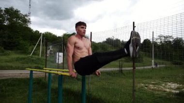 Güçlü bir adam karın kaslarını eğitir spor sahasında paralel barlarda bacak kaldırır. Genç üstsüz sporcu doğada egzersiz yapıyor. Doğada spor yapan sporcu. Sportif yaşam tarzı kavramı.