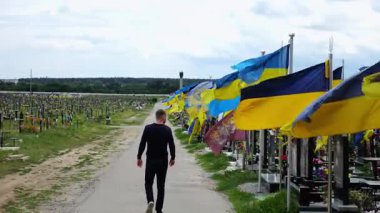 Man walking among countless graves of ukrainian soldiers with blue-yellow flags at military cemetery in Kharkiv. Mourning male standing near memorial pay tribute to fallen family member. Slow mo