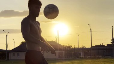 Genç adam gün batımında stadyumda futbol topuyla hokkabazlık yapıyor. Yeşil sahada profesyonel futbolcu topa vuruyor. Sporcu arka planda güneş ışığıyla çayırda numaralar yapıyor. Serbest stil futbol.