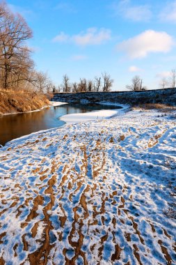 Güneşli bir kış günü, dondurucu nehrin ve yapraksız meşe ağaçlarının resimsel manzarası.