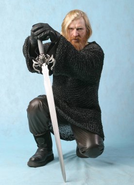 Sakallı, bıyıklı, zincirli zırhlı, arka planda hafif bir kılıçlı savaşçı portresi.