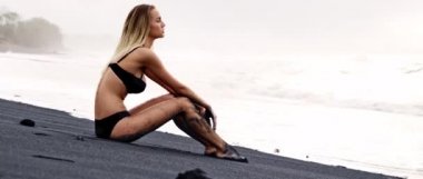 Koyu siyah kumsalda oturan bronz tenli genç kız ve okyanusun arka planına bak.