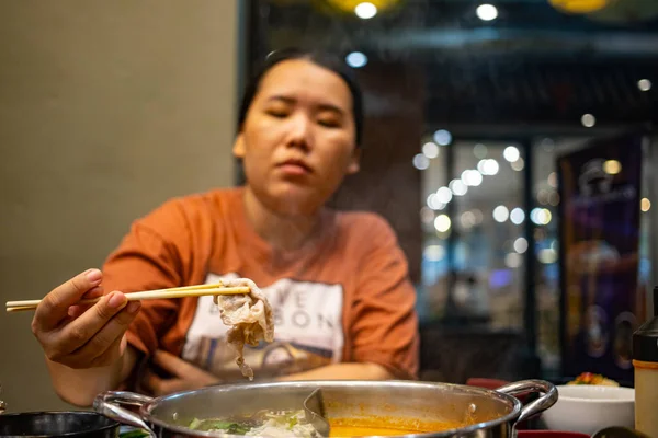 Mutlu tombul Asyalı kadın elinde bambu çubuklarıyla bir parça kaynamış biftekle Shabu restoranında yemek yiyor.