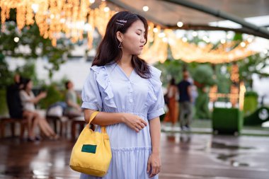 Açık mavi elbiseli güzel bir Asyalı kadın nehir kenarında duran küçük sarı bir çanta taşıyor.