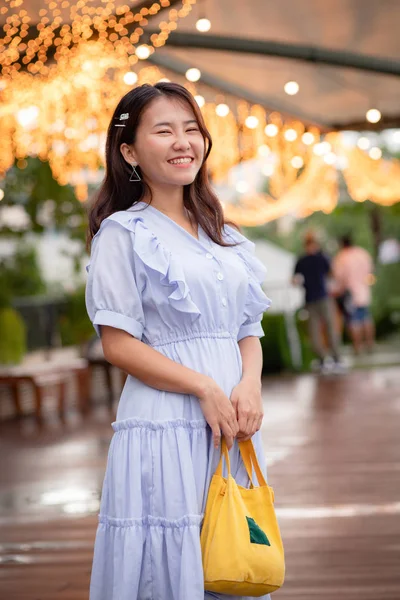 Happy adorable Asian woman in light blue dress carrying a small yellow tote bag who standing on the riverside with warm light decoration in background