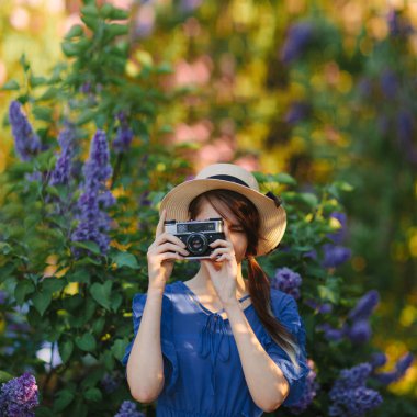 Retro kız, leylak çiçekli bahçede eski model fotoğraf makinesiyle fotoğraf çekiyor. Kare fotoğrafçılık.