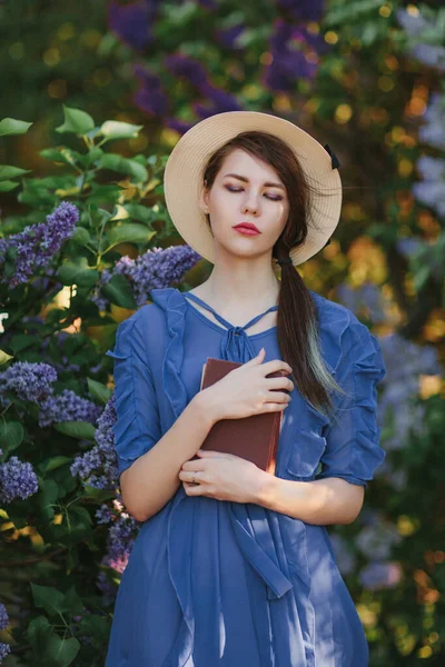 Genç güzel kız, çiçek açan bahçedeki leylak çalılarının yanında kitapla kal. Gözlerini kapadı, rüya gibi bir fotoğraf..