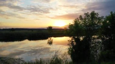 Gün batımında nehri gösteren hava aracı videosu. Renkli bulutlar, suda yansımalar ve manzaralı kırsal alan.