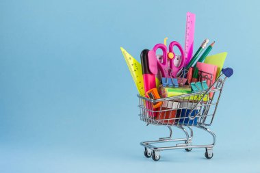 Açık mavi arka planda farklı kırtasiye malzemesi olan alışveriş arabası.