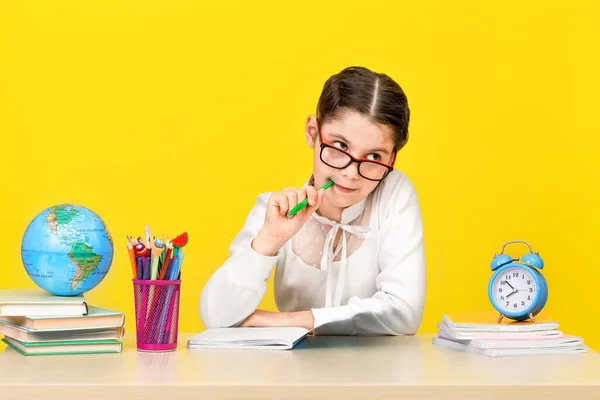 The schoolgirl sits at the desk and thinks about the decision of the task on yellow background.