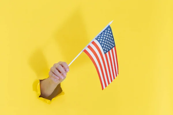 Hand aus einem Loch in gelbem Papier hält eine amerikanische Flagge mit blauen Sternen. lizenzfreie Stockfotos