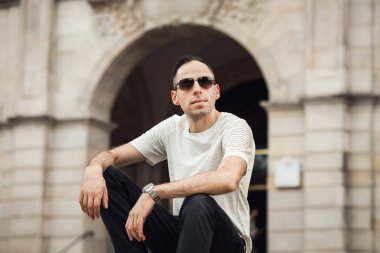 Adam gibi otur ve kameraya bak. Kahverengi gözlüklü DJ ve bina arka planında beyaz tişört.