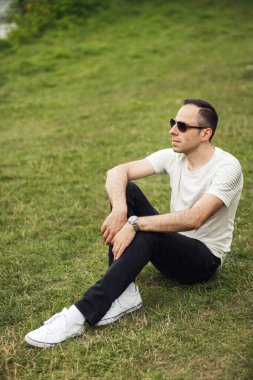 Gülümseyen bir adamın dikey fotoğrafı yeşil çimlerde oturur ve başka yere bakar. Beyaz tişörtlü DJ ve arka planda siyah pantolon.