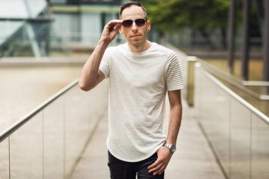 Şık giyimli bir adam şehirde dolaşıyor, kameraya bakıyor. Kahverengi gözlüklü DJ ve doğa arka planında beyaz tişört.