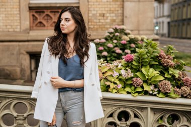 Beyaz moda ceketi ve kot pantolon giymiş gülümseyen çekici kadın başka tarafa bakıyor. Çiçekleri ve doğa geçmişi olan genç bir kadın.