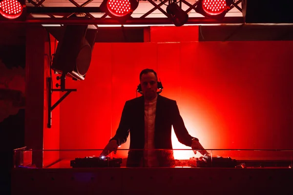 DJ with headphones at night club party. Man in black suit playing music on deck with vinyl record in red black interior