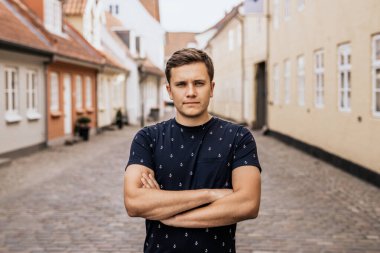 Kollarını kavuşturmuş ciddi bir adamın portresi. Şehrin arka planında koyu renk tişörtlü genç adam.