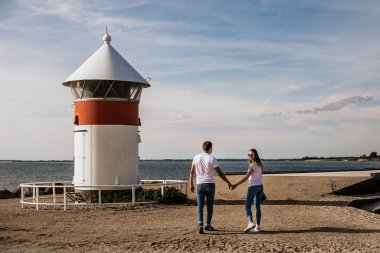 Mutlu romantik çift el ele tutuşup deniz feneriyle sahilde yürür. Seyahat yaşam tarzı konsepti
