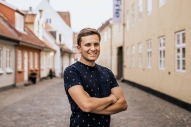 Kollarını kavuşturmuş gülümseyen adamın yan görüntüsü. Şehrin arka planında koyu renk tişörtlü genç adam.