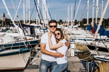 Deniz kıyısındaki mutlu genç çifte yakın durun. Beyaz tişörtlü, arka planda yelkenli bir çift.