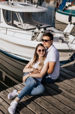 Deniz limanında oturan mutlu genç çiftin dikey fotoğrafı. Beyaz tişörtlü, yelkenli arka planda moda bir çift.