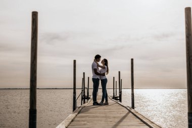 Romantik çiftin günbatımı liman manzarasının keyfini çıkarırken çekilmiş siluet fotoğrafı. Beyaz tişörtlü kadın ve erkek ambrance