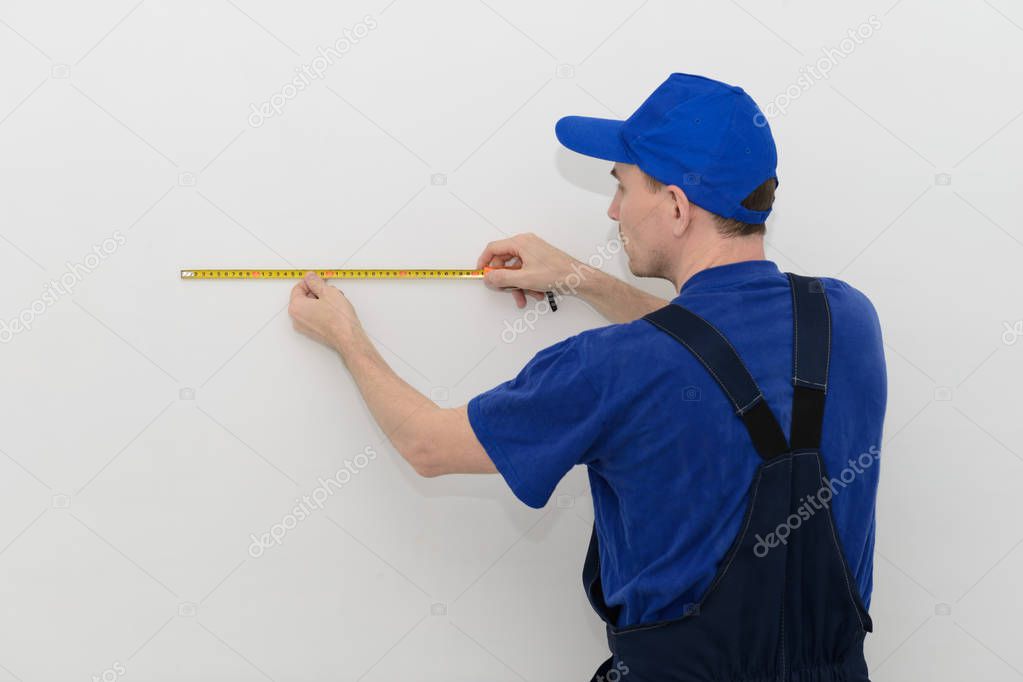 worker in blue overalls measures the distance on a white wall using a tape measure