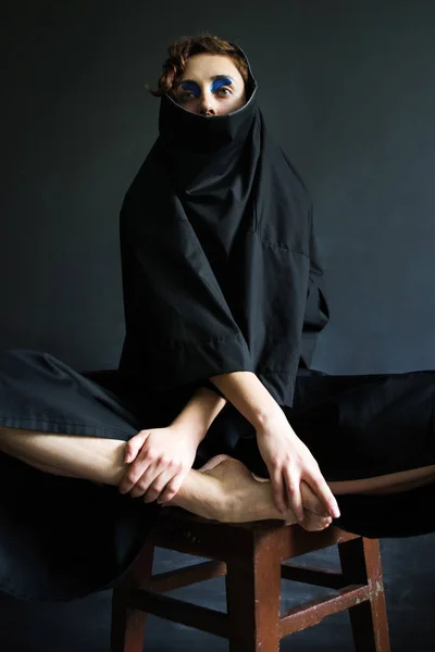 picture fashion photo girl with curly hair in voluminous black clothes with blue paint in front of her eyes in a hood sitting on a stool on a dark background hands and feet joined together