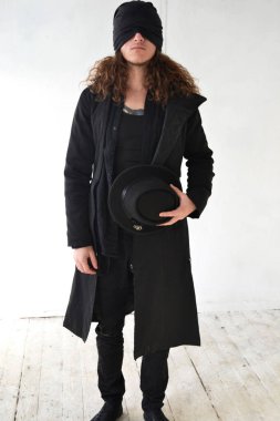 uzun kıvırcık saçlı siyah giysili bir şapka ile göz bağını elinde tutan