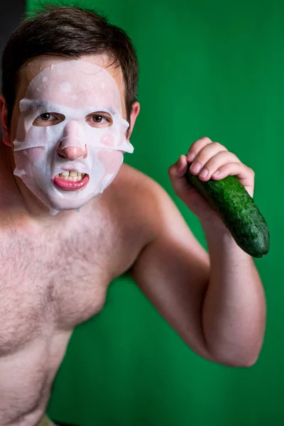 large man in a cosmetic face mask holds a green cucumber in his hand photo green background