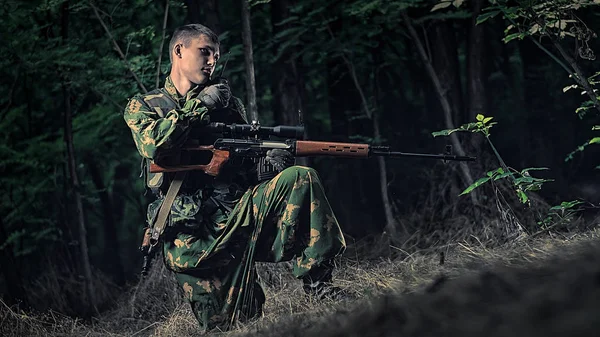 armed man in camouflage clothes in the forest with a sniper rifle speaks on a walkie-talkie