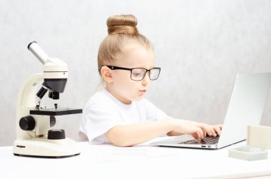 Beyaz arka planda, beyaz bir masada siyah gözlüklü küçük sarışın kız, dizüstü bilgisayarda çalışıyor. Masanın üzerinde bir mikroskop var..
