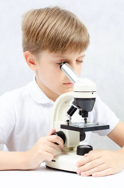 A blond-haired boy looking through a microscope, sitting at a white table, on a white background, vertical photo. — ストック写真