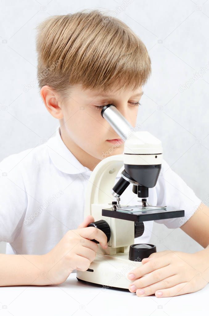 A blond-haired boy looking through a microscope, sitting at a white table, on a white background, vertical photo.