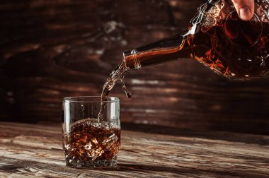 Viski ya da konyak güzel bir şişeden bir bardağa dökülür. Buz gibi hareket eden bir bardağa, doğal yaşlanmış ahşaptan yapılmış bir masa üstü, barlar için konsept, kahverengi ılık tonlar.