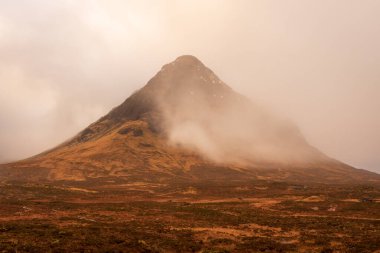 Kışlık kahverengi çimen tarlaları, İskoç dağlarında bulutlarla kaplı Bidean Nam Bian dağını kaplayan alçak asılı bulutlara bakar..